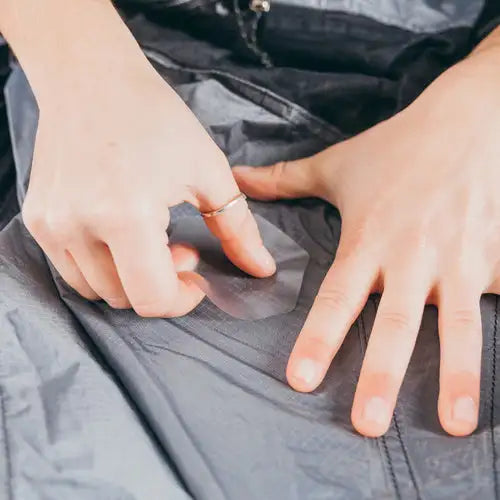A person applies a clear Gear Aid Tenacious Tape Hex Patch to a tear in gray fabric, possibly repairing outdoor gear. One hand holds the weatherproof patch while the other smooths the fabric.