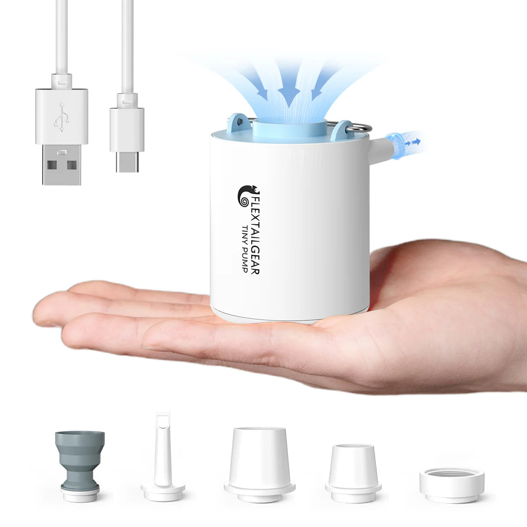 A compact white Flextail Tiny Pump Electric Air Pump rests on an open hand, emitting blue mist. Perfect for outdoor use, it comes with a USB charging cable and five nozzles—ideal as a rechargeable mini air pump for fast inflation.