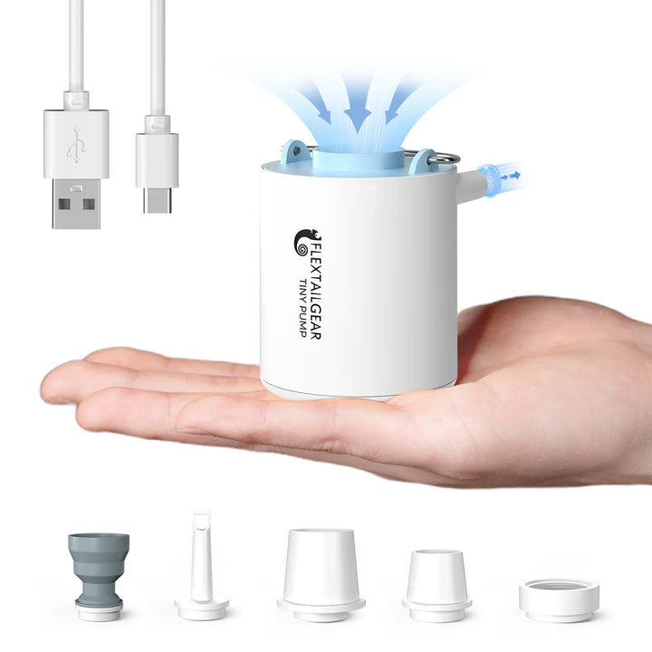 A compact white Flextail Tiny Pump Electric Air Pump rests on an open hand, emitting blue mist. Perfect for outdoor use, it comes with a USB charging cable and five nozzles—ideal as a rechargeable mini air pump for fast inflation.